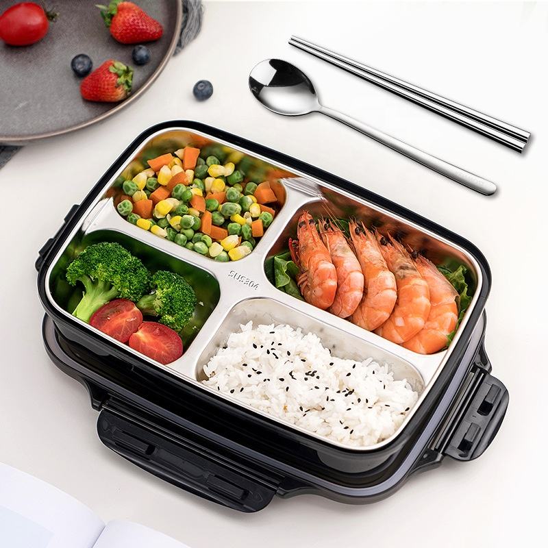 Black bento box with compartments filled with rice, vegetables, and shrimp, accompanied by a spoon and chopsticks on a white surface.