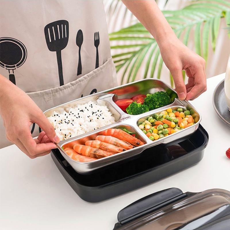 Person holding a stainless steel bento box with compartments of food on a white surface.